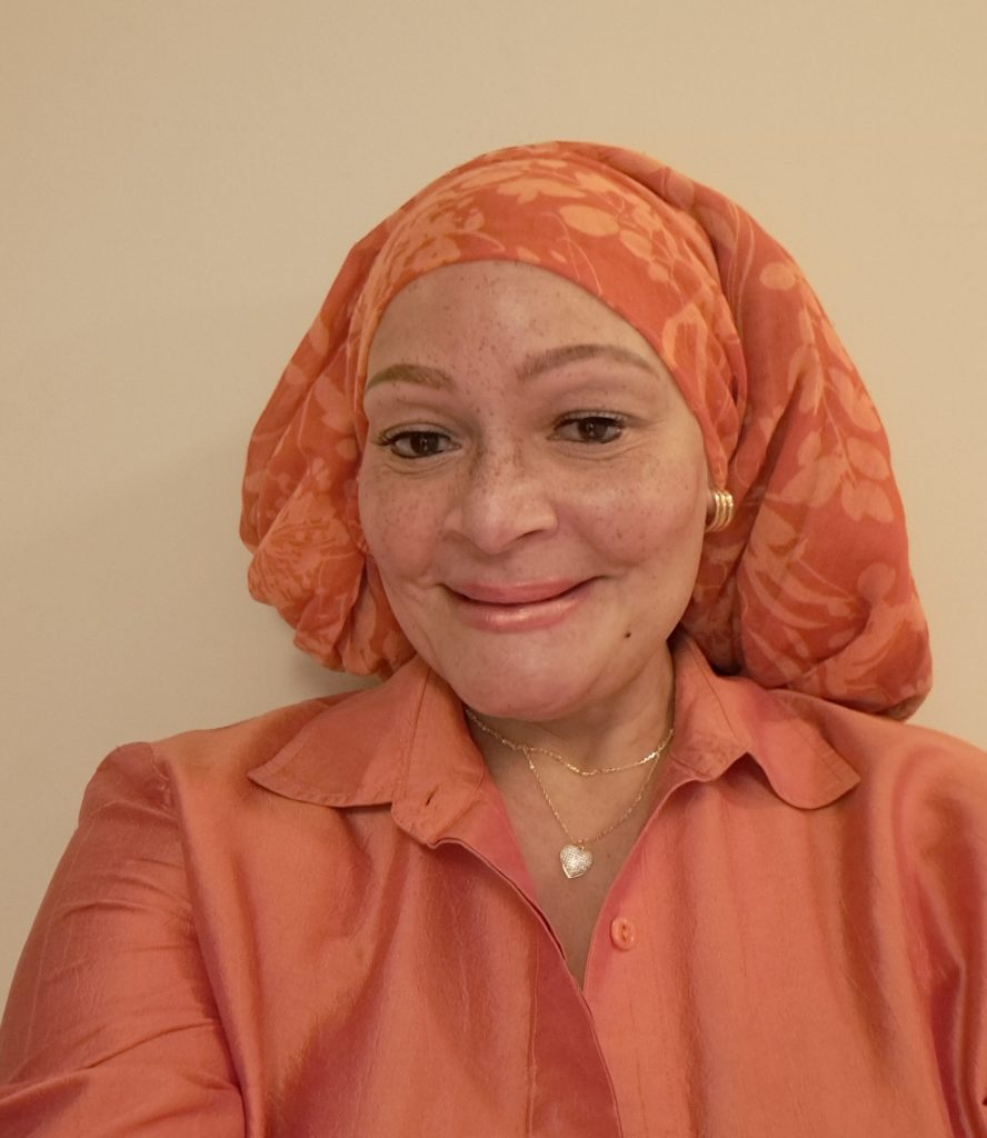 A woman with a bright orange headscarf and matching blouse smiles warmly at the camera. The background is a neutral color, highlighting her cheerful expression.